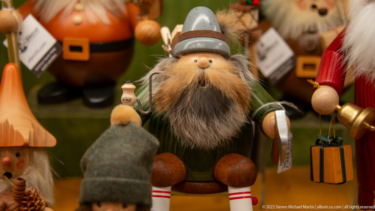 Bearded wooden holiday figurine in a gray pointed hat and green coat, holding small parcels among other craft figures.