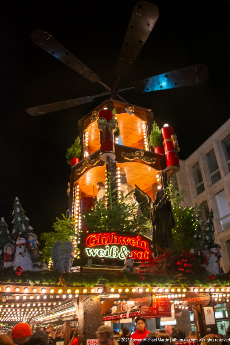 Illuminated Christmas market stall advertising Glühwein and Würstchen beneath a multi-tiered carousel-like roof decoration.