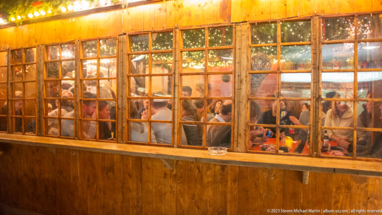 Diners seated inside a warmly lit wooden Christmas market hut, seen through rows of small-paned windows.
