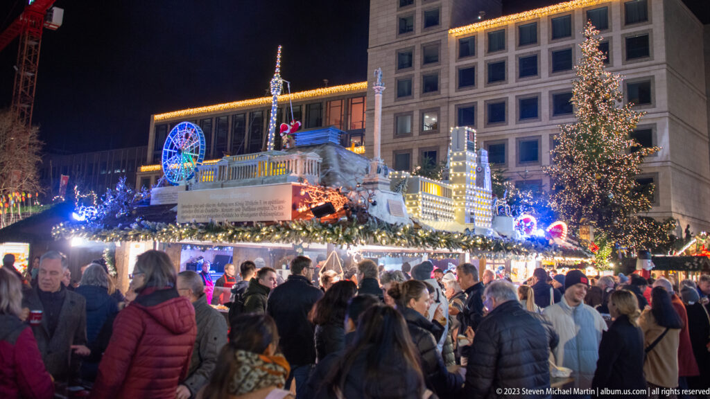 Large nighttime crowd moving between illuminated Christmas market stalls beneath a tall decorated tree in central Stuttgart.