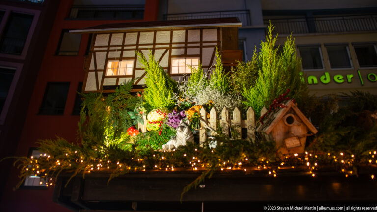 Miniature half-timbered house, fence, evergreens, flowers, lights, and birdhouse arranged on a Christmas market stall roof.
