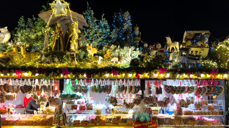 Illuminated Stuttgart Christmas-market food stall with hanging sausages below a roof crowded with miniature houses, figures, animals, and greenery.