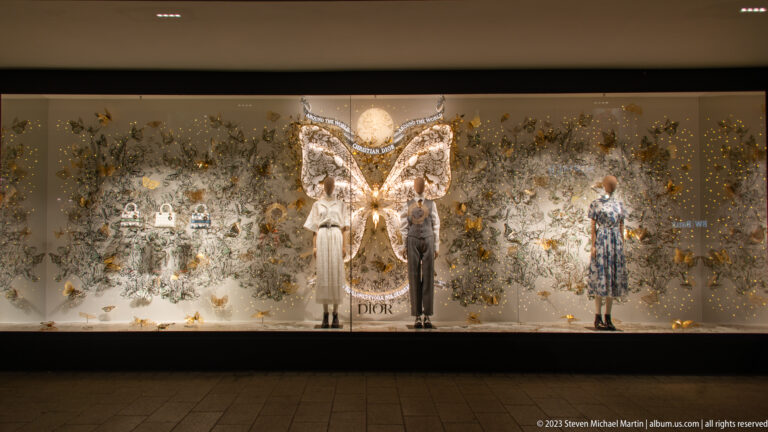 Dior store window with mannequins, metallic foliage, holiday lights, and a large glowing winged centerpiece.