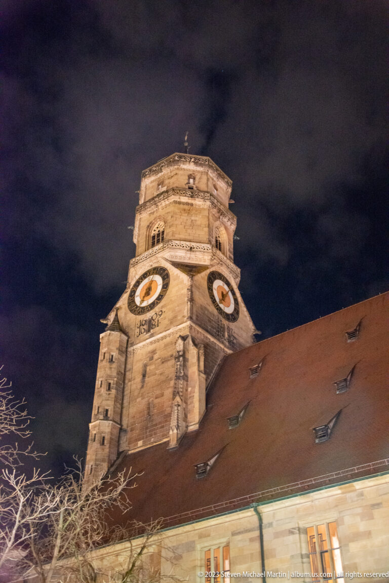Floodlit Stiftskirche clock tower rising above a steep red-tiled roof beneath a dark, clouded sky.