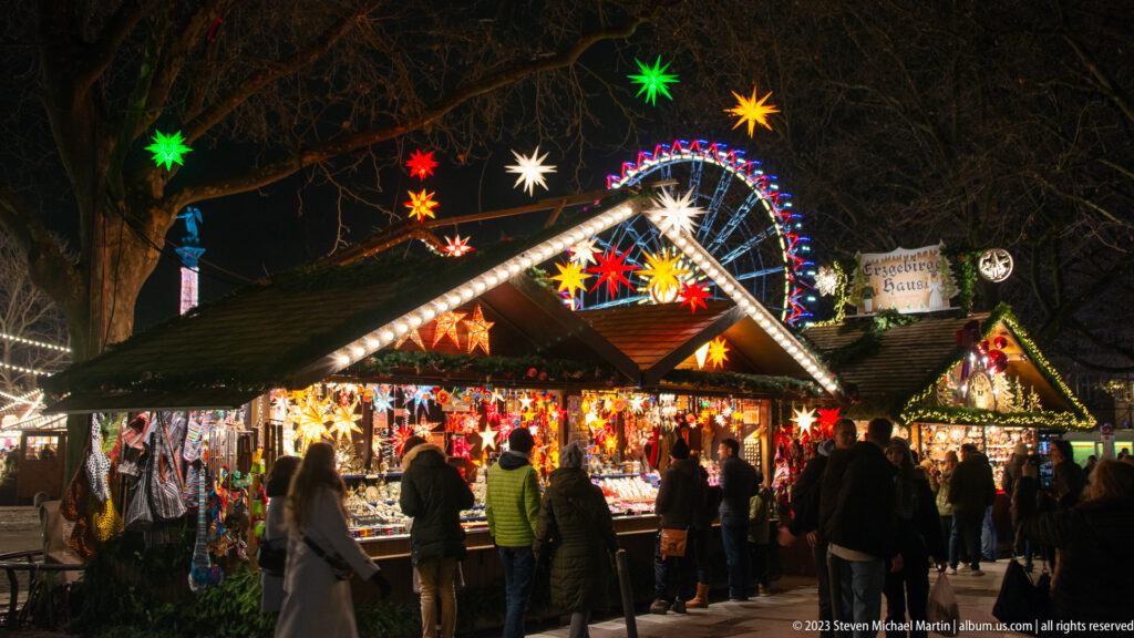 Crowds browse illuminated Christmas market stalls under colorful stars, with a Ferris wheel visible behind the trees.