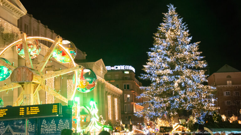 Tall Christmas tree densely covered in white lights beside a brightly illuminated fairground attraction in central Stuttgart.