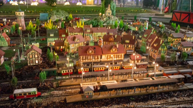 Miniature railway town with trains, station platforms, half-timbered buildings, streets, and landscaped scenery viewed from above.