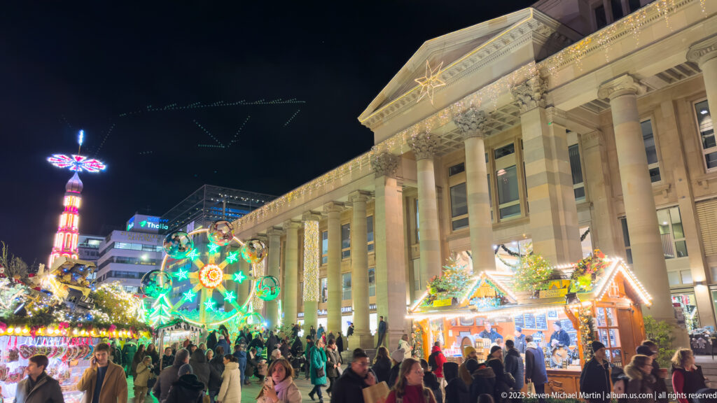 Crowded Christmas-market stalls in front of the long columned Königsbau on Stuttgart Schlossplatz at night.