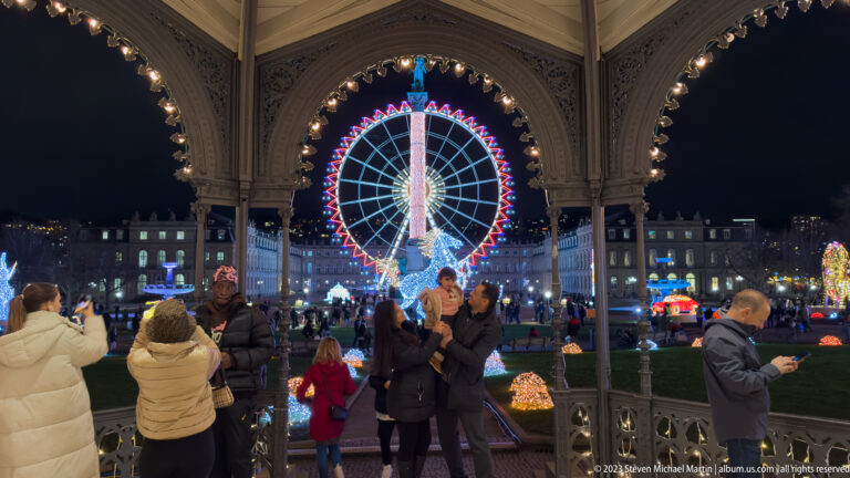 Visitors under the ornate Schlossplatz music pavilion, with a brightly lit Ferris wheel centered through the arches beyond.