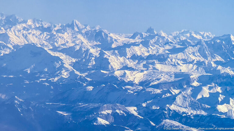 Snow-covered Alpine ridges and valleys seen from above beneath a clear blue sky.