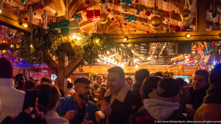 Crowd socializing inside a decorated timber market bar under ornaments, greenery, menus, and colored lights.