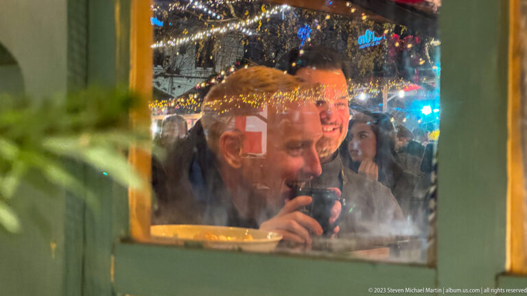 Man drinking from a dark cup behind a reflective window framed in green, with holiday lights reflected in the glass.