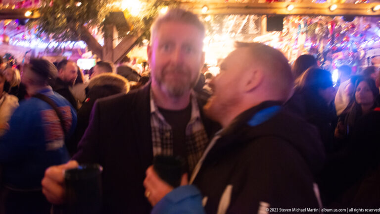Two men holding dark cups and talking closely in a crowded, brightly decorated market bar.