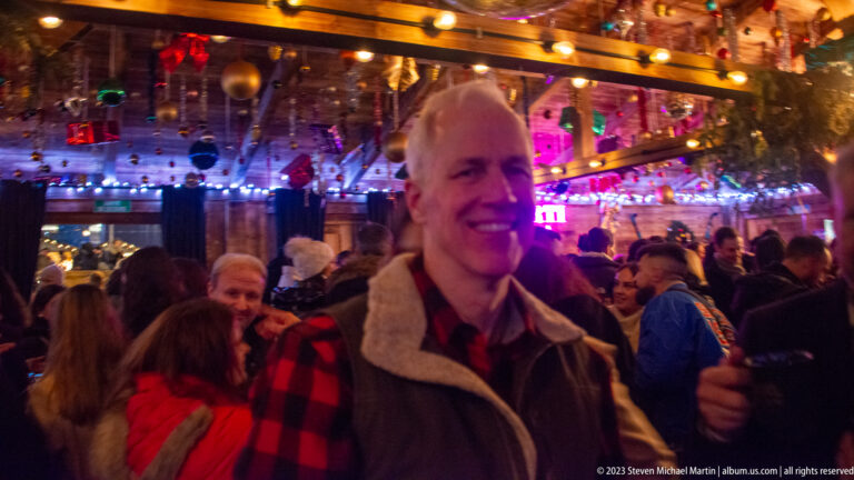 Smiling man in a checked shirt holding a dark cup inside a crowded, ornamented market pavilion.