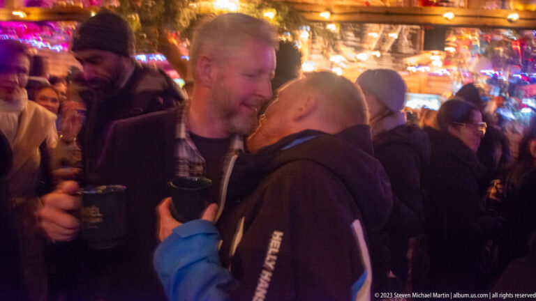 Two men holding cups lean toward each other in conversation amid a crowded, warmly lit market pavilion.
