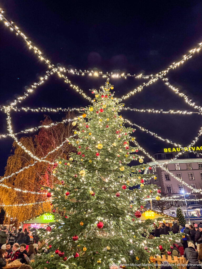 Tall decorated Christmas tree beneath radiating strands of white lights, with crowds and buildings around the square.