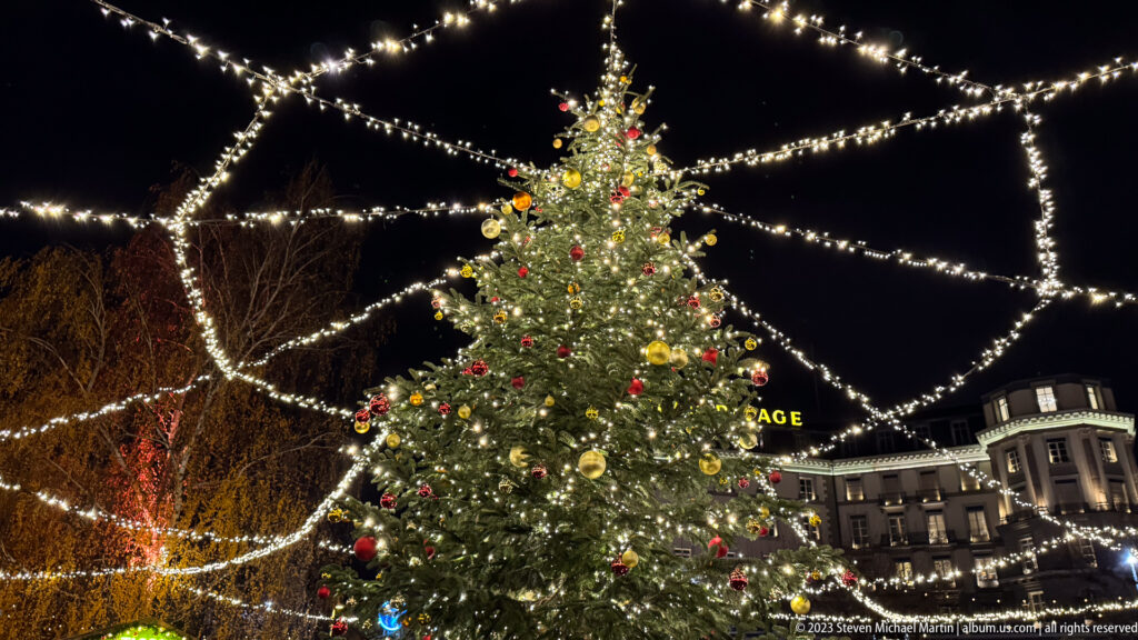 Decorated evergreen Christmas tree centered beneath a web of overhead string lights in an urban square at night.