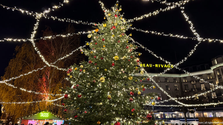 Illuminated Christmas tree with red and gold ornaments, overhead light strands, and BEAU-RIVAGE signage behind.