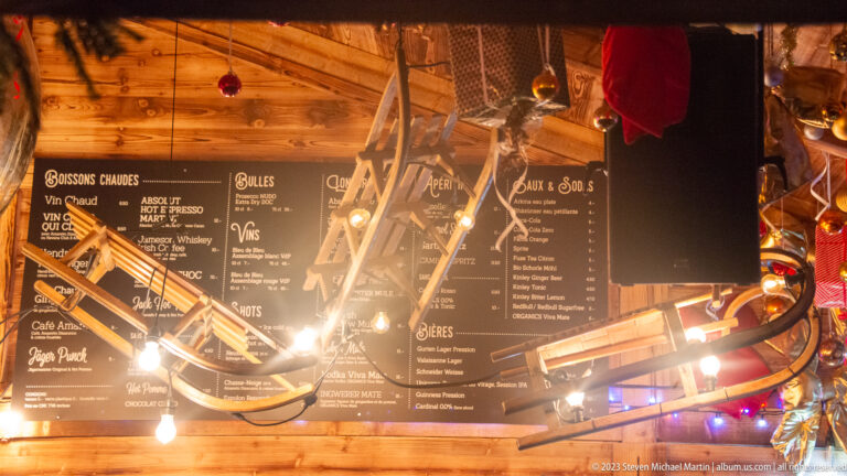 Black drink menu boards on a wooden market-bar wall behind hanging lamps, ornaments, and suspended equipment.