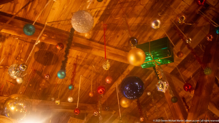 Red, blue, gold, silver, and turquoise baubles with a green gift ornament hanging from a timber ceiling.