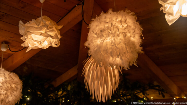 Feather-like white pendant lamp hanging from timber rafters among folded paper-style lights.