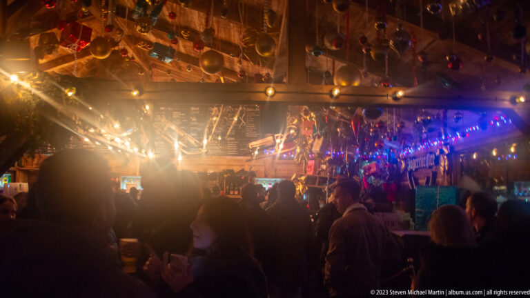 Crowded wooden market bar beneath hanging baubles and bright warm and colored lights at night.