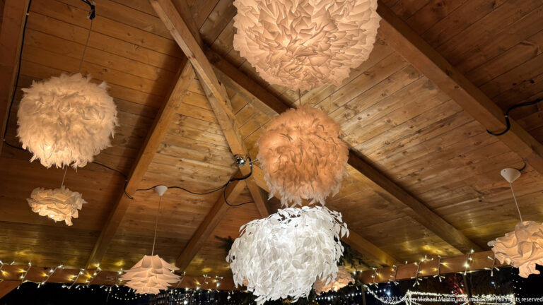 Multiple textured white pendant lamps hanging at different heights beneath a pitched wooden market roof.
