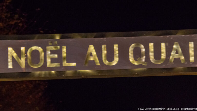 Close-up of illuminated gold letters reading “NOËL AU QUAI” on a dark sign at night.