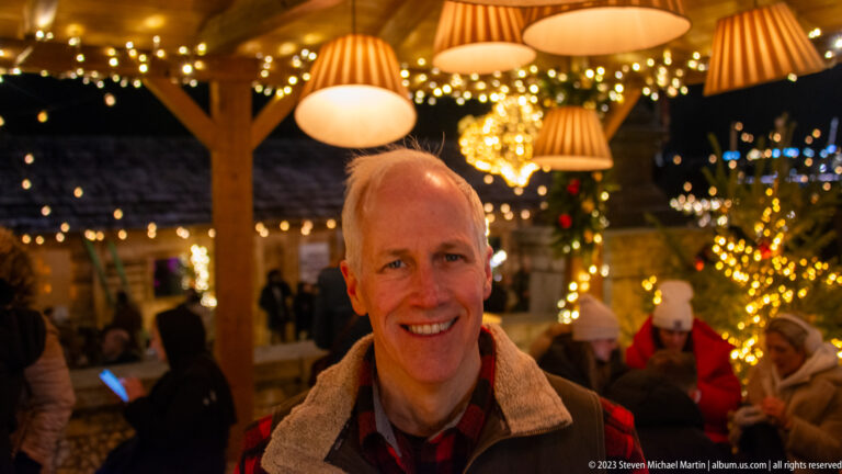Smiling man in a market pavilion beneath pleated lamps, fairy lights, greenery, and other visitors.