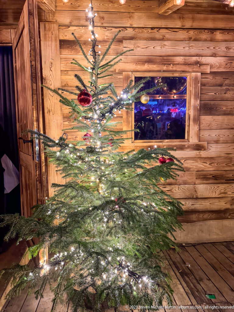 Small irregular evergreen decorated with white lights and red ornaments in front of a rustic wooden market wall.