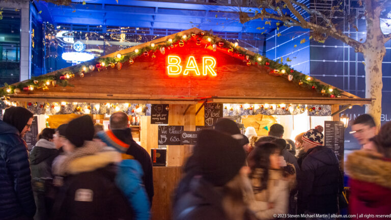 Wooden market chalet with a glowing BAR sign, garland, customers at the counter, and passing pedestrians.