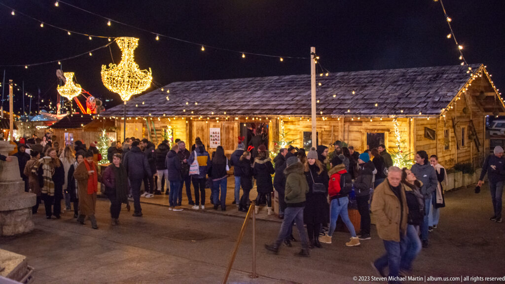 Crowd gathered beside illuminated wooden market chalets with chandelier-shaped holiday lights overhead.
