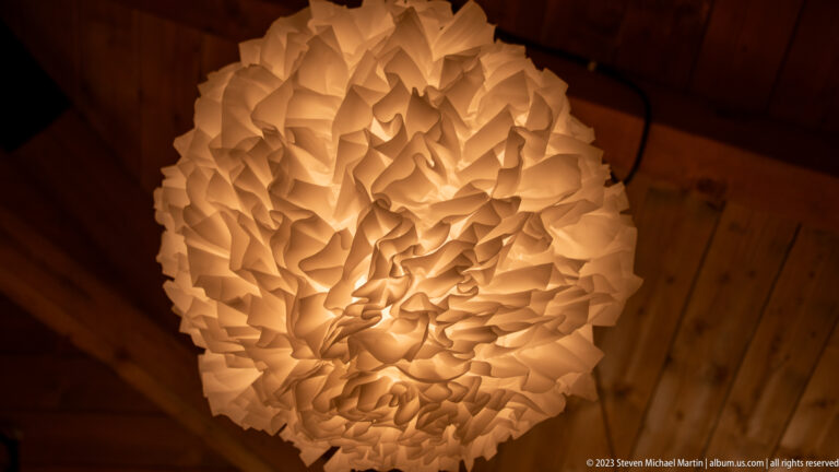 Glowing oval pendant shade made of densely layered white petal-like pieces beneath a wooden ceiling.