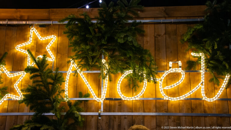 Warm white cursive “Noël” light sign with outlined stars and evergreen branches on a wooden wall.
