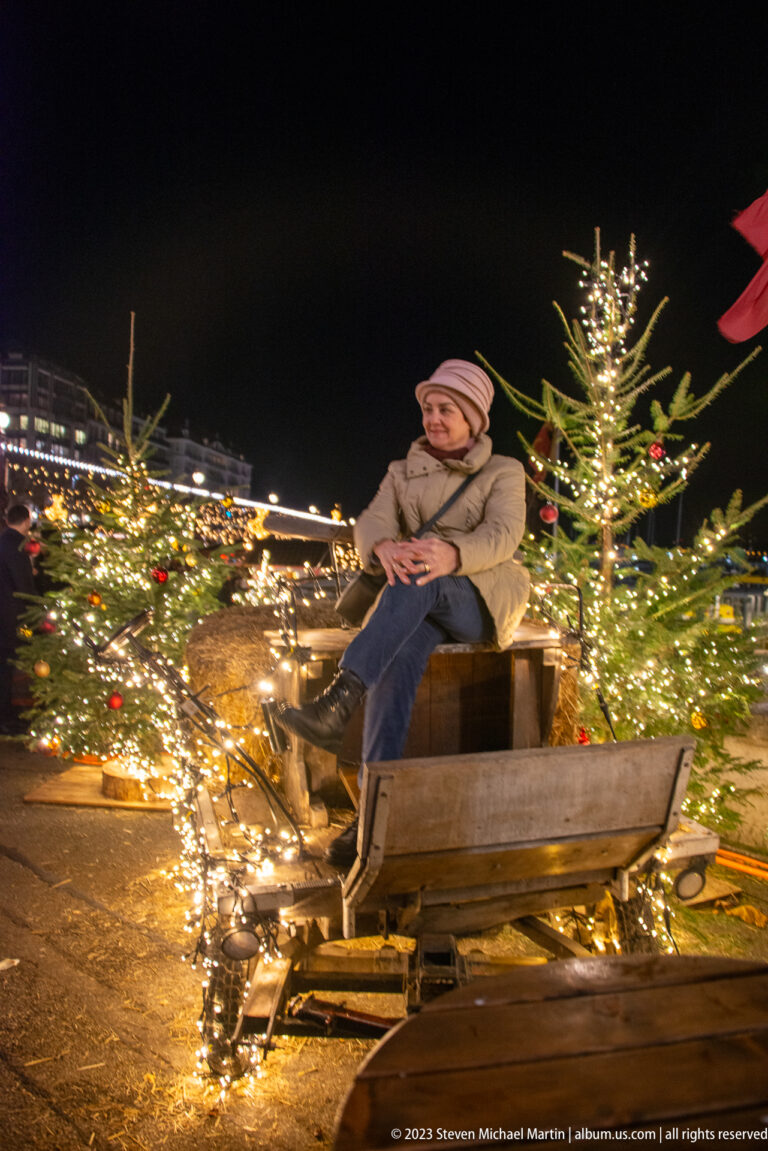 Bundled child seated on a rustic wooden sleigh among small fir trees covered with warm white lights.