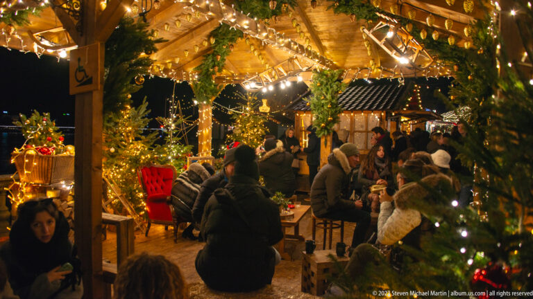 Visitors seated and standing inside a timber market pavilion decorated with greenery, fir trees, and warm lights.