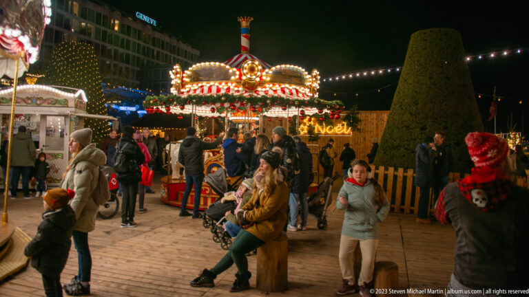 Families and visitors gather before a brightly illuminated carousel at the nighttime Christmas market.