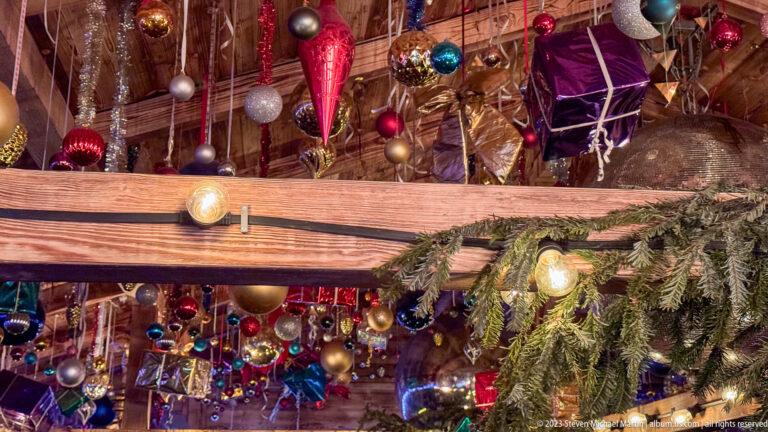 Colorful baubles and wrapped gift-box decorations densely suspended from timber rafters above evergreen branches.