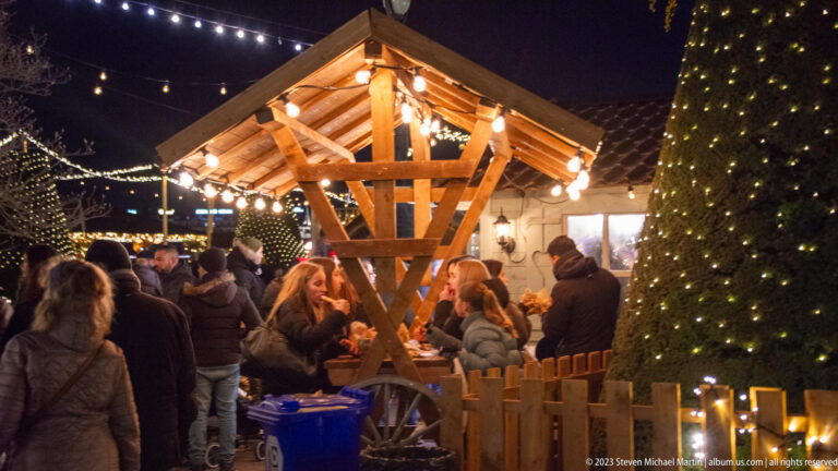 Visitors gather beside a lit peaked timber shelter and a tall clipped evergreen covered with tiny lights.