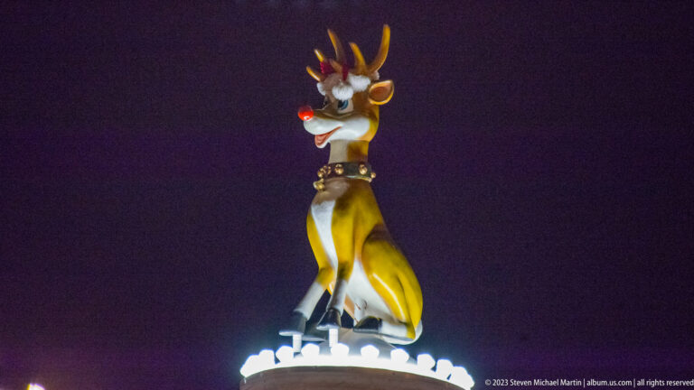 Large cartoon-like reindeer figure with antlers, red nose, and bell collar elevated against the night sky.