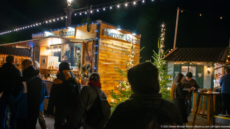 Customers queue at a lit wooden hot-drink chalet beside a small decorated fir tree.
