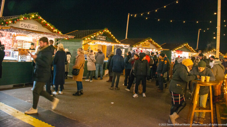 Crowds browse a row of brightly lit wooden market chalets decorated with garland and roofline lights.