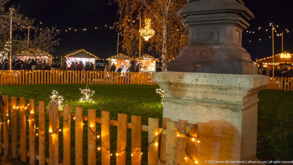 Light-wrapped wooden fence enclosing grass with small illuminated animal figures beside a large stone monument base.