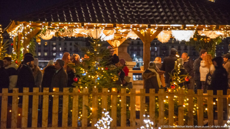 Lit small fir trees and a wooden fence in front of a crowded, garland-trimmed timber market pavilion.