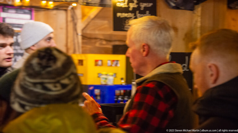 Winter-clad patrons gathered at a wooden market counter, with a gray-haired man in a checked shirt at center.