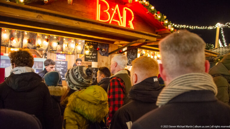Customers queue at a wooden market stall beneath a glowing red-orange BAR sign and festive garland.