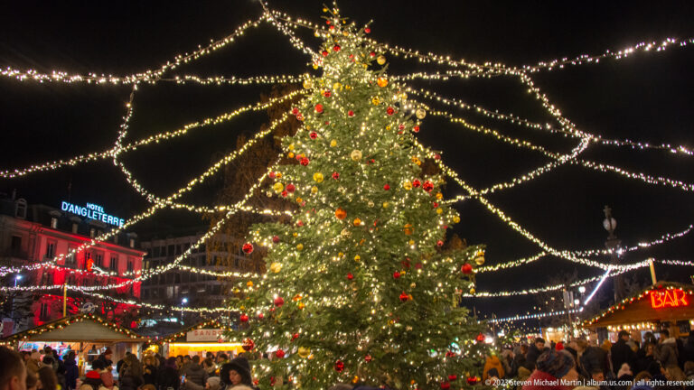 Large ornamented Christmas tree beneath radiating strands of lights above a crowded Geneva market square.