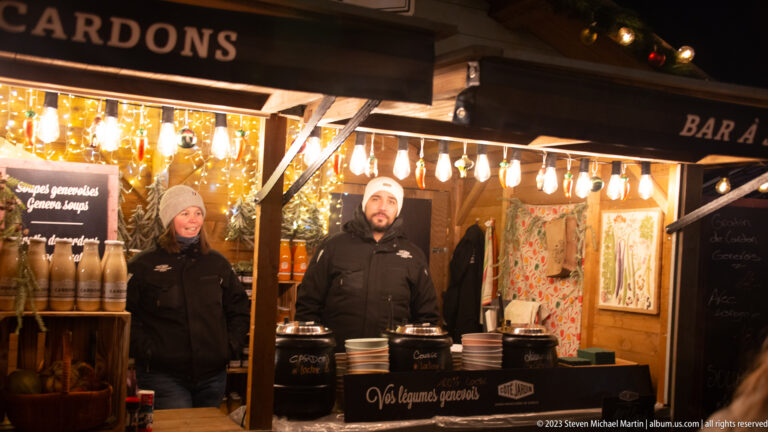 Two vendors behind a brightly lit wooden soup counter with black pots, bowls, bottles, and chalkboard menus.