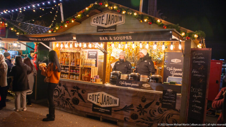 Wooden CÔTÉ JARDIN BAR À SOUPES stall with two vendors, soup pots, bottles, garland, and lights.