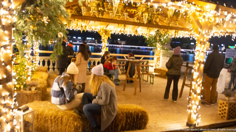 Visitors seated on hay bales inside a timber pavilion wrapped in warm lights and evergreen garlands.